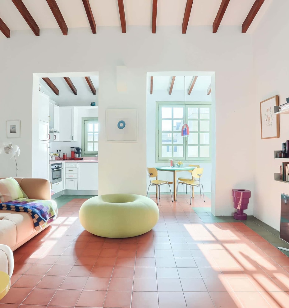 A bright living area featuring terracotta floor tiles, exposed wooden ceiling beams, a sofa with a striped cushion, and a round lime green ottoman, looking through to a dining area.