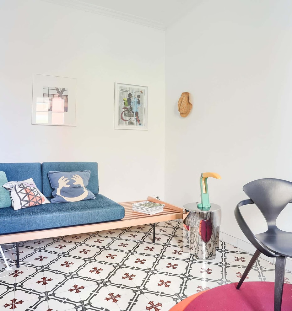 A bright living space featuring a blue sofa, a dark designer chair, and traditional patterned floor tiles.