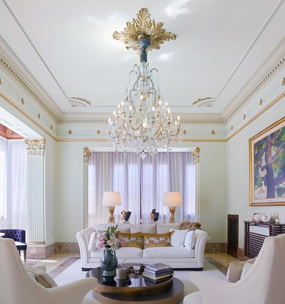 A formal living room with high ceilings, decorative gilded molding, and a crystal chandelier above a white sofa.
